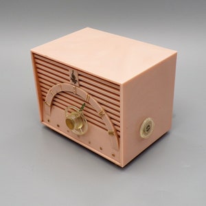K&ouml;nnte beinhalten: Ein vintage AM-Radio aus rosa Plastik mit einem goldenen Zifferblatt und einem Drehknopf. Das Radio hat ein "Emerson"-Logo auf der Vorderseite.