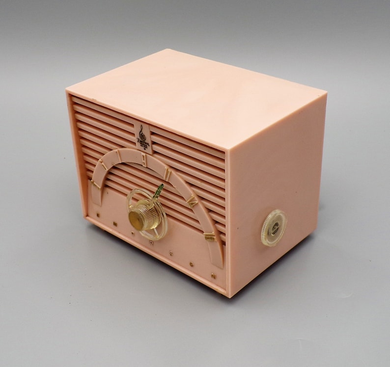 K&ouml;nnte beinhalten: Ein vintage AM-Radio aus rosa Plastik mit einem goldenen Zifferblatt und einem Drehknopf. Das Radio hat ein "Emerson"-Logo auf der Vorderseite.