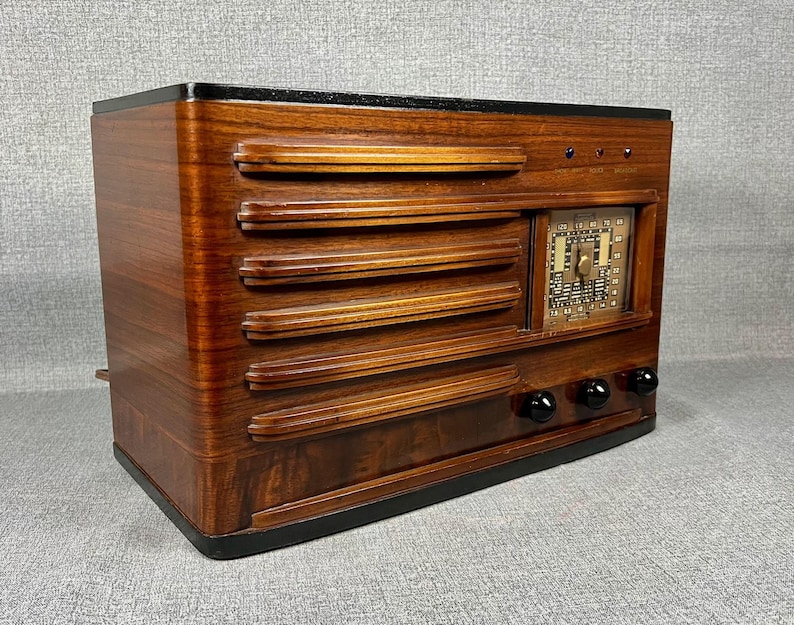 May include: A vintage wooden radio with a dark finish. The radio features horizontal slats across the front, a square dial, and three black knobs. The top and base are black, and the overall design is rectangular.
