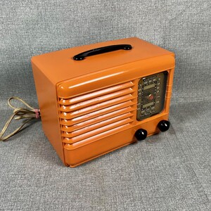 Puede incluir: Una radio naranja vintage con asa y perillas negras. La radio tiene una rejilla de altavoz y un dial de sintonizaci&oacute;n. El cable de alimentaci&oacute;n es visible en el lado izquierdo. Dispositivo electr&oacute;nico de estilo retro.