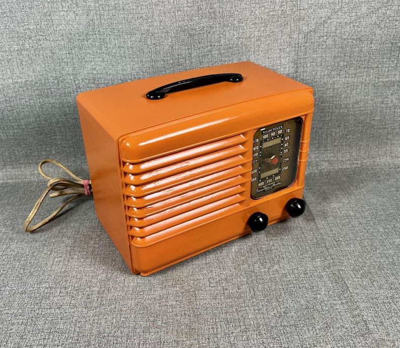 Puede incluir: Una radio naranja vintage con asa y perillas negras. La radio tiene una rejilla de altavoz y un dial de sintonizaci&oacute;n. El cable de alimentaci&oacute;n es visible en el lado izquierdo. Dispositivo electr&oacute;nico de estilo retro.