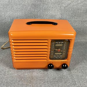 Puede incluir: Radio vintage naranja con asa y perillas negras. La radio tiene un dial con n&uacute;meros y una ventana transparente. Tiene una rejilla de altavoz y un cable de alimentaci&oacute;n.