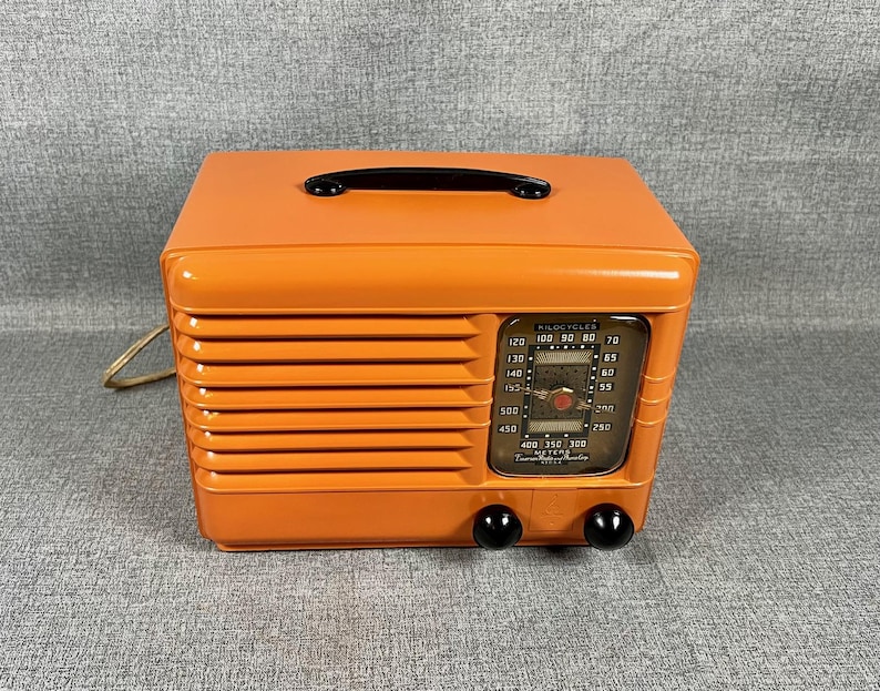 Puede incluir: Radio vintage naranja con asa y perillas negras. La radio tiene un dial con n&uacute;meros y una ventana transparente. Tiene una rejilla de altavoz y un cable de alimentaci&oacute;n.