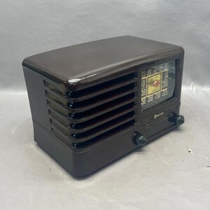 K&ouml;nnte beinhalten: Ein vintage braunes Plastikradio mit einem silbernen Zifferblatt und einem schwarzen Drehknopf. Das Radio hat eine Holzmaserung und ein Metallgitter.