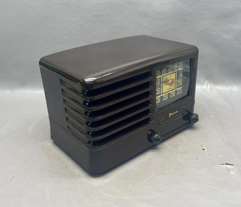 K&ouml;nnte beinhalten: Ein vintage braunes Plastikradio mit einem silbernen Zifferblatt und einem schwarzen Drehknopf. Das Radio hat eine Holzmaserung und ein Metallgitter.