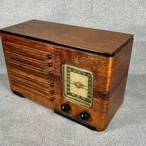 May include: A vintage wooden radio with a rectangular design. The radio has a dark wood finish, a dial with numbers, and two black knobs. The front has a speaker grill design. The radio is a classic piece of electronics.