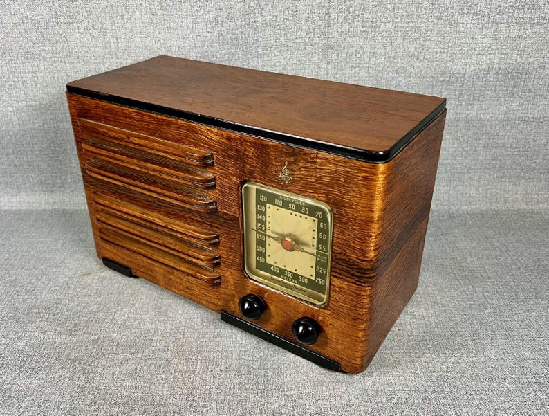 May include: A vintage wooden radio with a rectangular design. The radio has a dark wood finish, a dial with numbers, and two black knobs. The front has a speaker grill design. The radio is a classic piece of electronics.