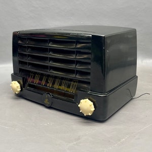 Op de afbeelding: Een zwarte vintage radio met een witte wijzerplaat en knoppen. De radio heeft een metalen rooster en een houten kast.