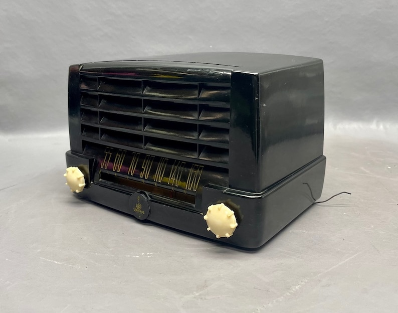 Op de afbeelding: Een zwarte vintage radio met een witte wijzerplaat en knoppen. De radio heeft een metalen rooster en een houten kast.
