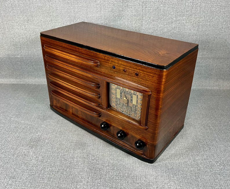 May include: A vintage wooden radio with a dark brown finish. The radio has a rectangular shape with a speaker grill on the front and a dial display. It features black knobs and a glossy top.