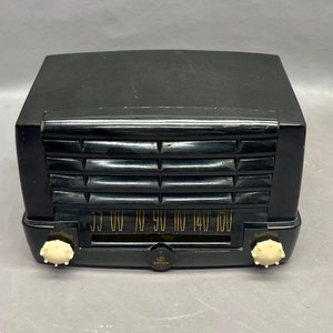 Op de afbeelding: Een zwarte vintage Zenith radio met een witte wijzerplaat en knoppen. De radio heeft een houtnerf afwerking en een metalen rooster. De wijzerplaat heeft nummers van 55 tot 160.
