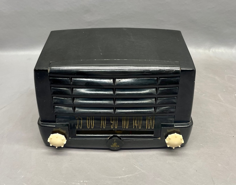 Op de afbeelding: Een zwarte vintage Zenith radio met een witte wijzerplaat en knoppen. De radio heeft een houtnerf afwerking en een metalen rooster. De wijzerplaat heeft nummers van 55 tot 160.