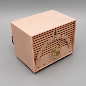 K&ouml;nnte beinhalten: Ein vintage Radio aus rosa Plastik mit einem goldfarbenen Zifferblatt und einem Drehknopf. Das Radio hat ein "Philco" Logo und ein Musiknotensymbol.