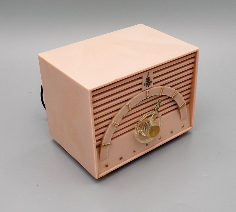 K&ouml;nnte beinhalten: Ein vintage Radio aus rosa Plastik mit einem goldfarbenen Zifferblatt und einem Drehknopf. Das Radio hat ein "Philco" Logo und ein Musiknotensymbol.