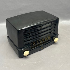 Op de afbeelding: Een zwarte vintage radio met een witte wijzerplaat en knoppen. De radio heeft een zwarte kunststof behuizing en een metalen rooster.