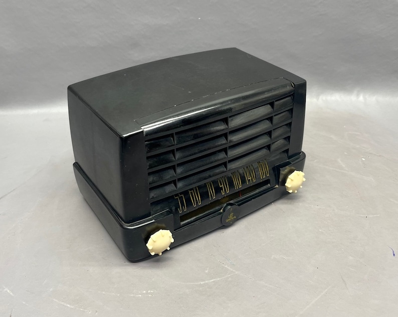 Op de afbeelding: Een zwarte vintage radio met een witte wijzerplaat en knoppen. De radio heeft een zwarte kunststof behuizing en een metalen rooster.