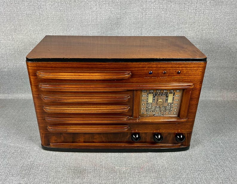 May include: A vintage wooden radio with a dark brown finish. The radio features a speaker grill design on the front, a tuning dial, and several knobs. The radio's design is reminiscent of mid-century modern aesthetics.