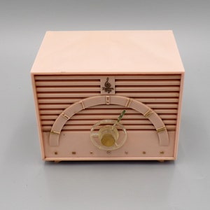 K&ouml;nnte beinhalten: Ein rosafarbenes Emerson-Radio im Vintage-Stil mit einem goldfarbenen Drehregler und einem goldfarbenen H&ouml;henregler. Das Radio hat ein goldfarbenes Logo mit einem Violinschl&uuml;ssel und dem Text "Emerson" und "Supertone".