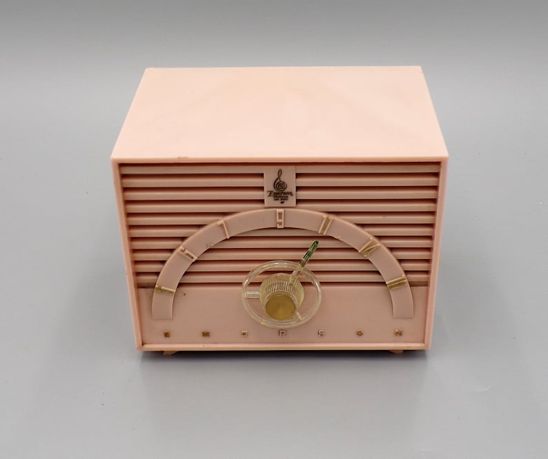 K&ouml;nnte beinhalten: Ein rosafarbenes Emerson-Radio im Vintage-Stil mit einem goldfarbenen Drehregler und einem goldfarbenen H&ouml;henregler. Das Radio hat ein goldfarbenes Logo mit einem Violinschl&uuml;ssel und dem Text "Emerson" und "Supertone".