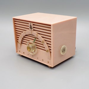 K&ouml;nnte beinhalten: Ein vintage-Radio von Emerson in rosa mit einem goldenen Zifferblatt und einem Drehknopf. Das Radio hat ein Holzmaserungsmuster und das Wort "Emerson" ist auf der Vorderseite aufgedruckt.