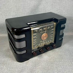 May include: A black Crosley radio with a glossy finish, featuring a central glass dial with gold-toned text and a copper-colored circular knob. The radio has silver-colored mesh speaker grilles on both sides and three dark brown control knobs.