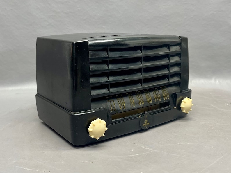 Op de afbeelding: Een zwarte vintage radio met een witte wijzerplaat en knoppen. De radio heeft een zwarte kunststof behuizing en een metalen rooster.