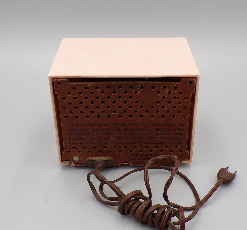 K&ouml;nnte beinhalten: Ein rosafarbenes Vintage-Radio von Emerson mit einem braunen Lautsprechergitter und einem Warnhinweis mit dem Text: "THIS COVER IS EQUIPPED WITH AN INTERLOCK TO PREVENT DANGEROUS ELECTRICAL SHOCK FOR SERVICING, REMOVE SCREWS PULL COVER COMPLETELY OUT Op от ZAMPER TH INTERLOCK PLUG OR ITS SAFE OF FEATURE WILL BE DESTROYED EMIRSON RADIO PHONOGRAPH  MADE IN U.S.A."