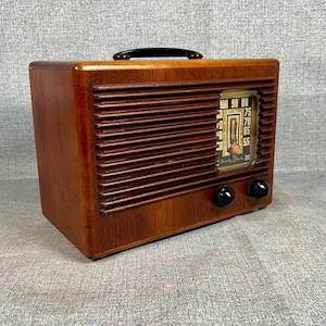 May include: A rectangular wooden radio with a dark brown finish, featuring horizontal slats across the front, a black carrying handle on top, and two black circular knobs. The radio has a glass-covered dial with numbers and a small orange indicator.