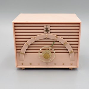 K&ouml;nnte beinhalten: Ein rosafarbenes Emerson-Radio im Vintage-Stil mit einem goldfarbenen Drehregler und einem goldfarbenen Logo mit der Aufschrift "Emerson Radio and Phonograph Corp."