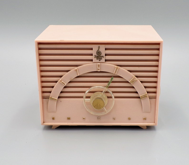 K&ouml;nnte beinhalten: Ein rosafarbenes Emerson-Radio im Vintage-Stil mit einem goldfarbenen Drehregler und einem goldfarbenen Logo mit der Aufschrift "Emerson Radio and Phonograph Corp."
