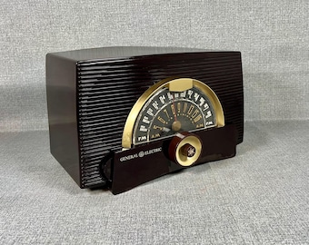 May include: A vintage General Electric radio in a dark brown, rectangular cabinet with horizontal ribbed detailing. The radio features a gold-toned, semi-circular dial with black numerals and a brown tuning knob. The text "GENERAL ELECTRIC" is visible.