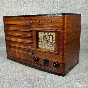 May include: A vintage wooden radio with a dark finish. The radio features a rectangular design with horizontal slats and a central dial. Three black knobs are visible on the bottom, and the dial displays numbers and a needle. The top has the words "SHORT WAVE POLICE BROADCAST".