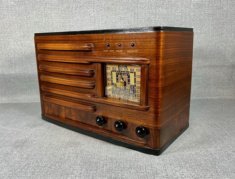 May include: A vintage wooden radio with a dark finish. The radio features a rectangular design with horizontal slats and a central dial. Three black knobs are visible on the bottom, and the dial displays numbers and a needle. The top has the words "SHORT WAVE POLICE BROADCAST".