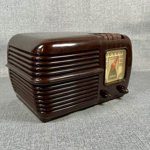 May include: A vintage brown radio with a glossy finish. The radio has a rectangular shape with horizontal lines on the sides and a dial with numbers and a needle. Two knobs are visible on the front.
