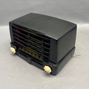 Op de afbeelding: Een zwarte vintage radio met een witte wijzerplaat en knoppen. De radio heeft een rooster aan de voorkant en een snoer dat uit de achterkant komt.