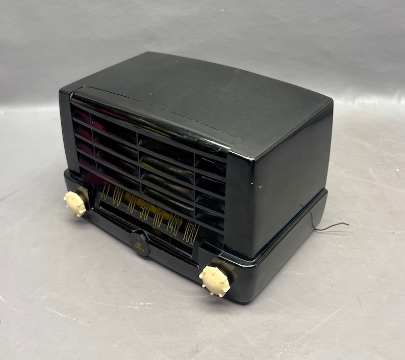 Op de afbeelding: Een zwarte vintage radio met een witte wijzerplaat en knoppen. De radio heeft een rooster aan de voorkant en een snoer dat uit de achterkant komt.