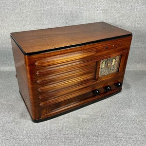 May include: A vintage wooden radio with a glossy, dark brown finish. The radio features horizontal, ridged detailing on the front and a rectangular display with a dial. Three black knobs are located below the display.