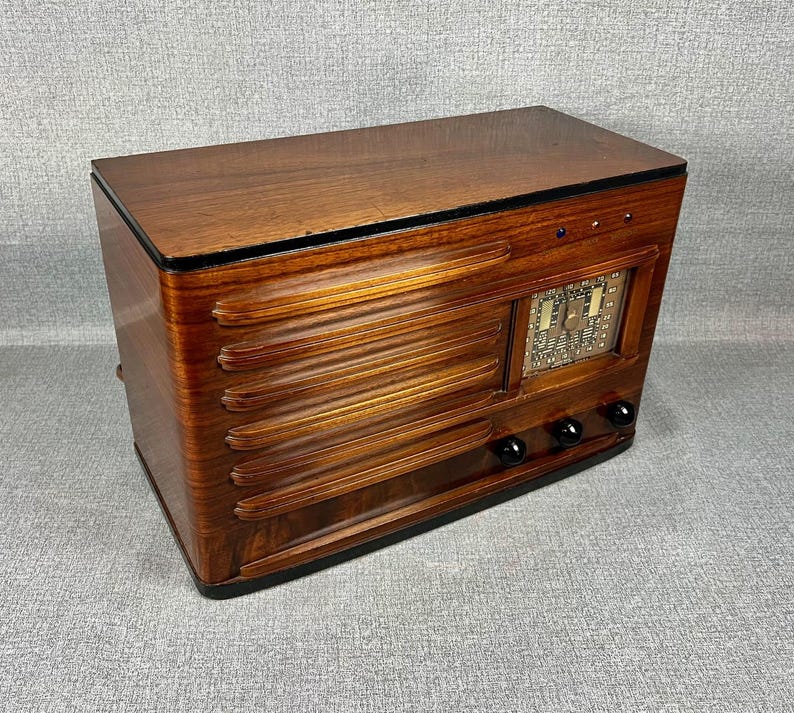 May include: A vintage wooden radio with a glossy, dark brown finish. The radio features horizontal, ridged detailing on the front and a rectangular display with a dial. Three black knobs are located below the display.
