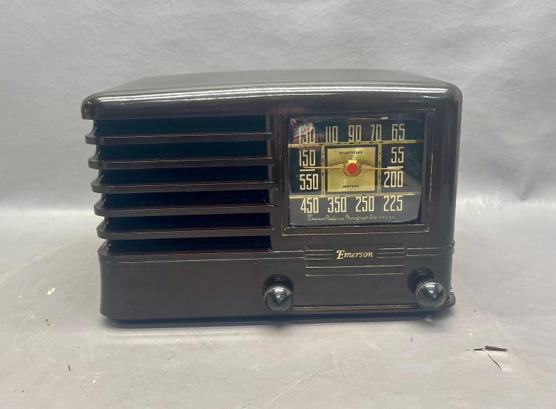 K&ouml;nnte beinhalten: Ein braunes Vintage-Radio von Emerson mit einem schwarzen Zifferblatt und einer goldfarbenen Tuningskala. Das Radio hat ein Lautsprechergitter an der Vorderseite und einen Knopf an jeder Seite.
