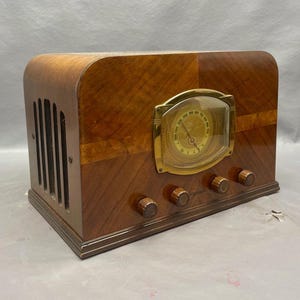 May include: A vintage wooden radio with a gold-colored dial and knobs. The radio has a wood grain pattern and a speaker grille on the side.