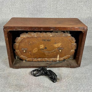 May include: A vintage wooden clock radio in a dark brown finish. The clock face is oval and visible through the front. A black power cord is coiled in front of the radio.