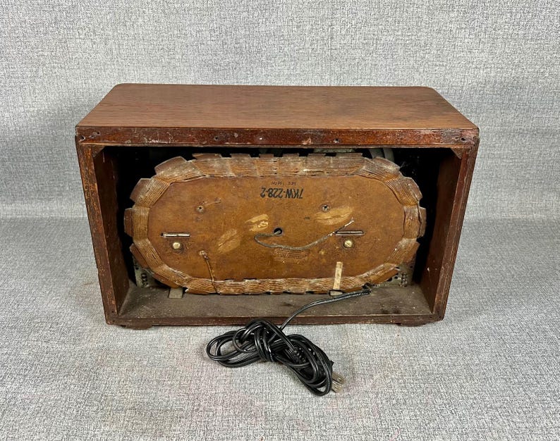 May include: A vintage wooden clock radio in a dark brown finish. The clock face is oval and visible through the front. A black power cord is coiled in front of the radio.