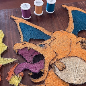 May include: A handmade string art piece featuring a Charizard design, with orange, blue, and purple thread on a wooden board. Three spools of thread in purple, tan, and blue are visible. The artwork depicts a popular character.