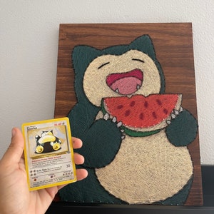 May include: String art of Snorlax eating watermelon, crafted on a wooden board. The artwork features green, beige, and red threads. A Snorlax Pokémon card is held in front of the artwork.