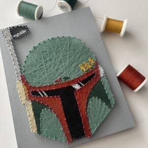 May include: String art depiction of a Mandalorian helmet on a gray wooden board. The helmet is crafted with green, red, black, and gold thread, with small pink pins outlining the design. Several spools of thread in various colors are scattered around the board.
