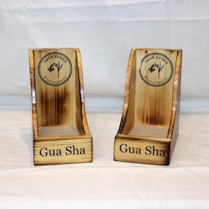 May include: Two wooden display stands with a curved design. The stands are made of light-colored wood with a darker, burnt-wood effect. Each stand has the text "Gua Sha" at the base and a circular logo that reads "Skin Linen" and "Mindful Beauty".