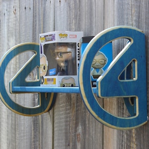 May include: A blue wooden shelf in the shape of the number four, with a gold trim. The shelf holds two Funko Pop figures. The shelf is mounted on a weathered wooden fence.