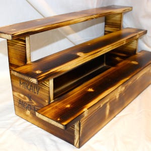 May include: A three-tiered wooden shelf with a rustic, burnt finish. The shelf is labeled "Mikaly & Aigo" and is perfect for displaying small items.