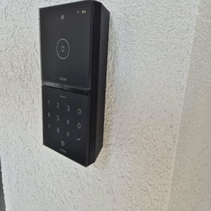 Op de afbeelding: Een zwart rechthoekig slim intercomsysteem met een camera en een deurbelknop op het bovenste gedeelte, en een numeriek toetsenbord op het onderste gedeelte. De merknaam Loxone staat in witte letters op zowel het bovenste als onderste paneel.