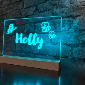 以下が含まれることがあります： 白いベースに青いLEDライト。ライトは白い蝶と蜂のデザインと、「Holly」という名前が筆記体で書かれています。